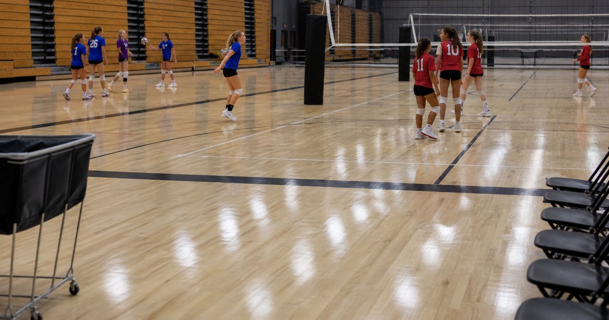 Half empty school gymnasium before a youth volleyball tournament, two small groups of young players in different colored jerseys warming up on one court while nets stand idle on the neighboring courts, empty wooden bleachers pulled out along the wall, bright overhead lights, no signage