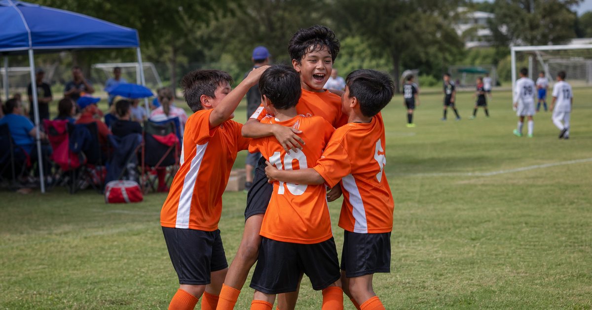 Youth soccer tournament on a sunny park field, a team in orange jerseys celebrating a goal on a side pitch while families watch from folding chairs under a canopy tent, another game visible on the main field in the background