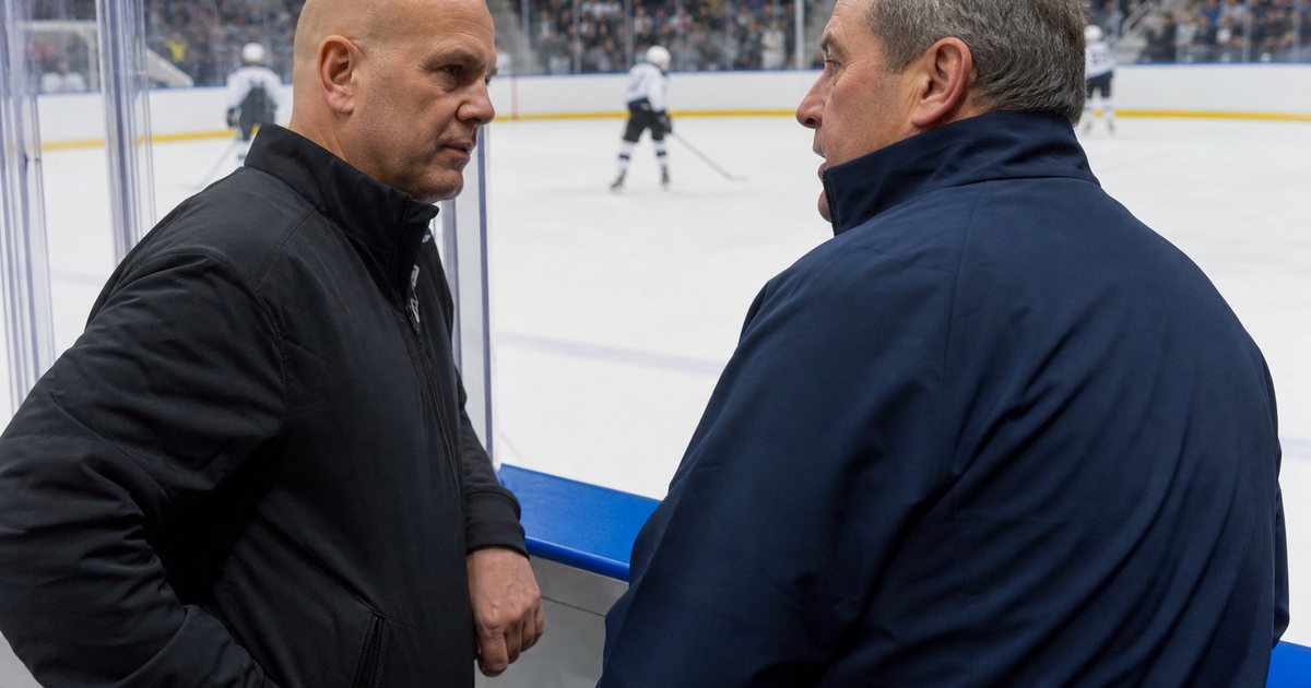 Two coaches in team jackets talking at the boards of an ice rink during a break in play, players skating in the background, arena lights overhead, spectators in the stands behind the glass