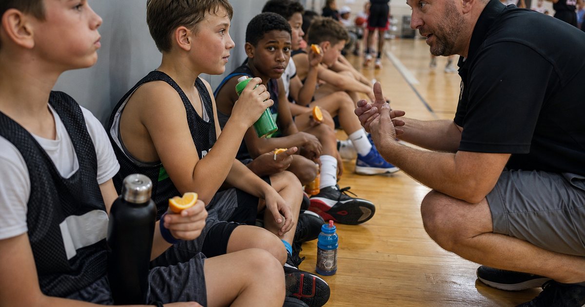 Youth basketball team sitting on a gym floor along the wall between games, players drinking from water bottles and eating orange slices, a coach crouched in front of them talking, another game in progress on the court behind