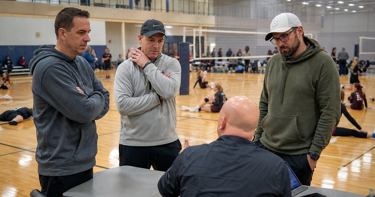 Youth volleyball tournament in a school gymnasium, three coaches gathered around a folding scorer's table talking with a tournament director, players stretching on the court behind them, bright overhead lights, no signage
