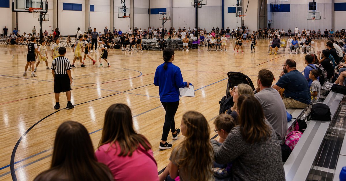 Wide view inside a large indoor sports complex with four basketball courts side by side, games in progress on each, referees in striped shirts, families on bleachers, a tournament staffer with a clipboard walking between the courts